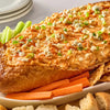 A creamy Buffalo chicken dip topped with blue cheese and green onions is served inside a hollowed-out bread Barrel, with carrots, celery, and bread pieces for dipping.