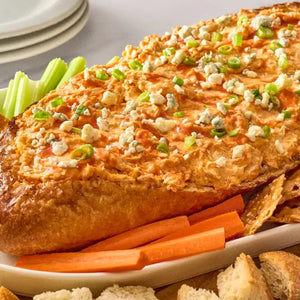 A creamy Buffalo chicken dip topped with blue cheese and green onions is served inside a hollowed-out bread Barrel, with carrots, celery, and bread pieces for dipping.