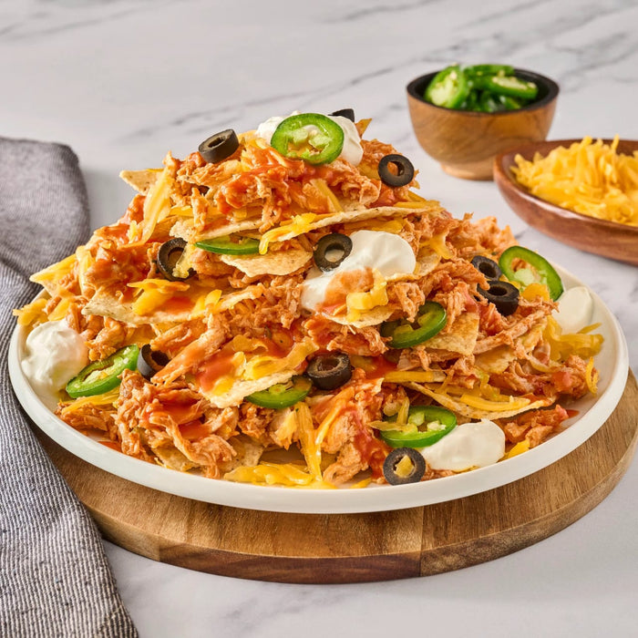 A large platter of chicken nachos is piled high with cheese, sour cream, olives, and jalapeños, with extra toppings served on the side in small wooden Barrel bowls.