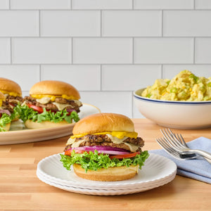 A juicy double cheeseburger sits on a white plate from the Barrel collection, with more burgers and a bowl of potato salad in the background.