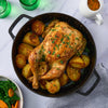 A whole roasted chicken and potatoes are served in a black cast-iron pan, accompanied by a side of vegetables and a gravy boat.