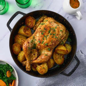 A whole roasted chicken and potatoes are served in a black cast-iron pan, accompanied by a side of vegetables and a gravy boat.