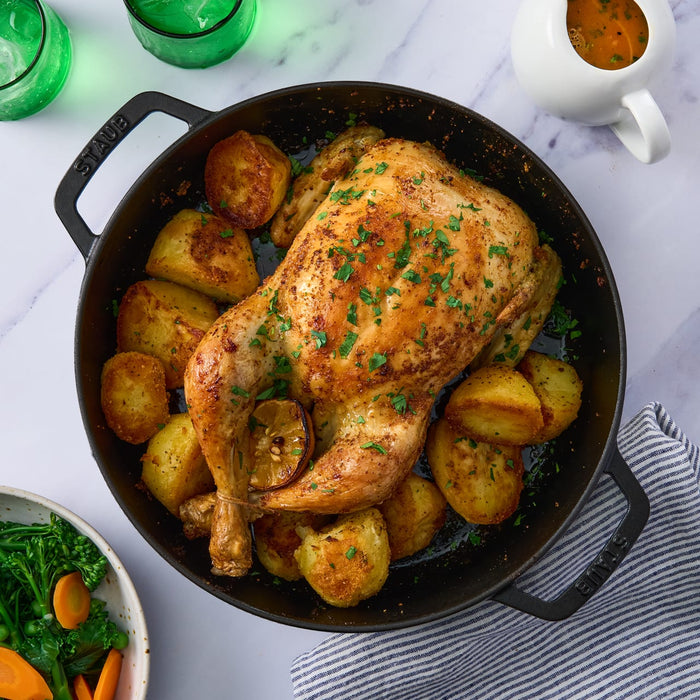 A whole roasted chicken and potatoes are served in a black cast-iron pan, accompanied by a side of vegetables and a gravy boat.