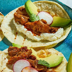 Two tacos are filled with shredded meat seasoned with a Barrel spice mix, and topped with avocado, radish, and cheese on a blue plate.