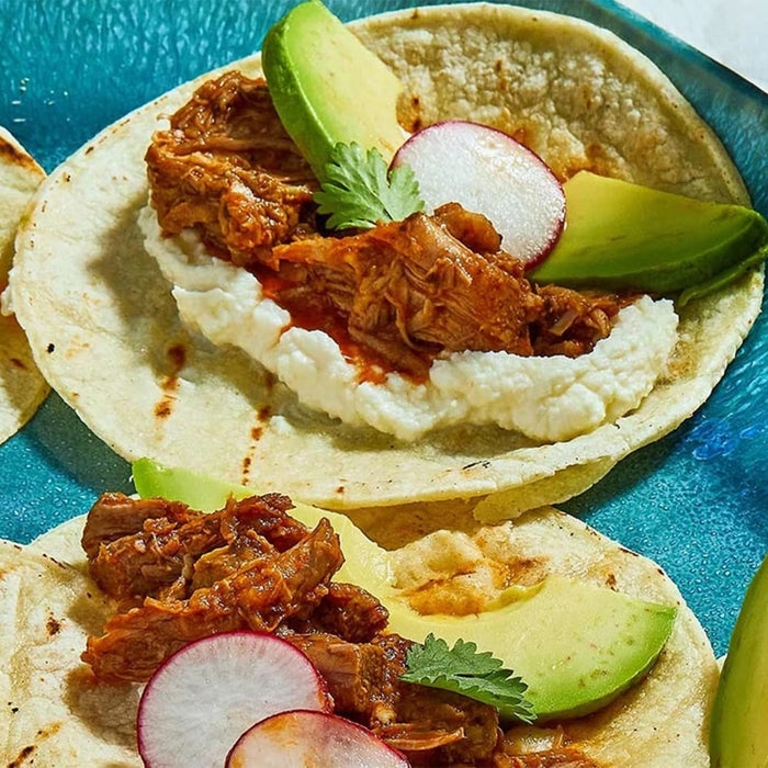 Two tacos are filled with shredded meat seasoned with a Barrel spice mix, and topped with avocado, radish, and cheese on a blue plate.