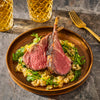 A plate of perfectly cooked lamb chops served over a bed of greens and grains, accompanied by golden beverages.