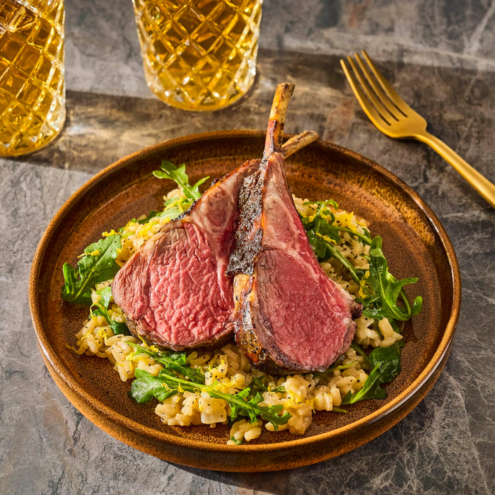 A plate of perfectly cooked lamb chops served over a bed of greens and grains, accompanied by golden beverages.