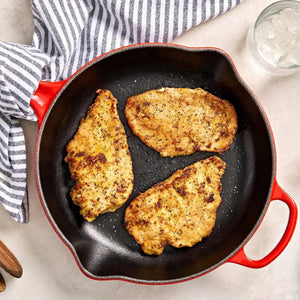 A top-down view shows three golden-brown, seasoned chicken cutlets cooking in a red and black cast iron Barrel skillet.