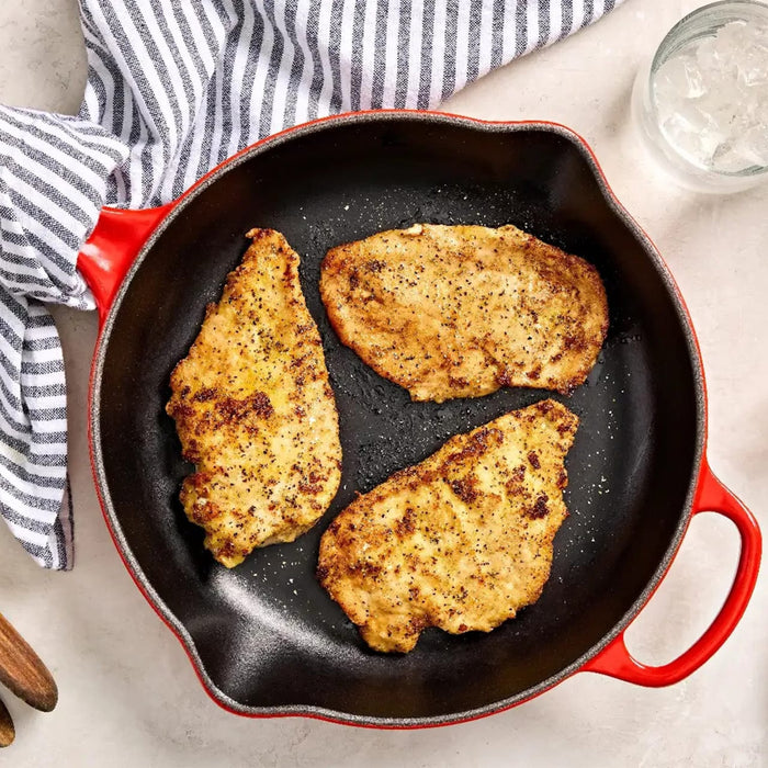 A top-down view shows three golden-brown, seasoned chicken cutlets cooking in a red and black cast iron Barrel skillet.