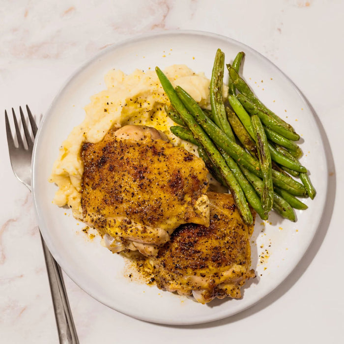 A delicious-looking meal on a white plate features two golden-brown chicken thighs seasoned with a Barrel spice blend, served alongside creamy mashed potatoes and sautéed green beans.