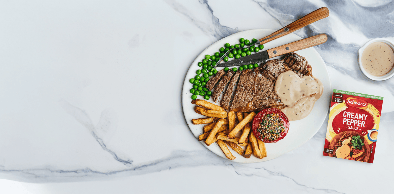 Steak with fries, peas, and a packet of creamy pepper sauce on a marble surface.