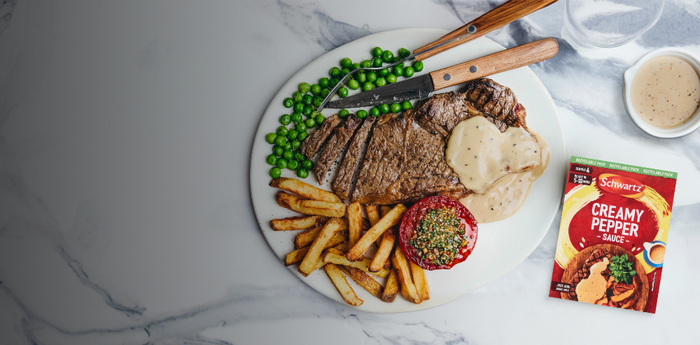 Steak with fries, peas, and a packet of Schwartz creamy white pepper sauce on a marble surface.