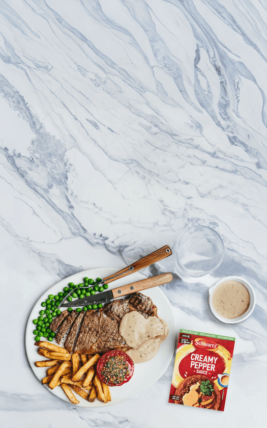 Steak with fries, peas, and a packet of creamy pepper sauce on a marble surface.