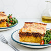 A hearty slice of a layered pasta casserole with a meat sauce and creamy béchamel topping, sprinkled with herbs, is served on a plate with a fresh arugula salad.