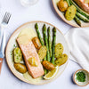 An overhead shot of a plated meal featuring a salmon fillet with lemon sauce, roasted asparagus, and small potatoes sprinkled with herbs.