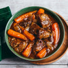 A hearty bowl of beef stew with large chunks of carrots is presented in a green bowl on a white wooden table.