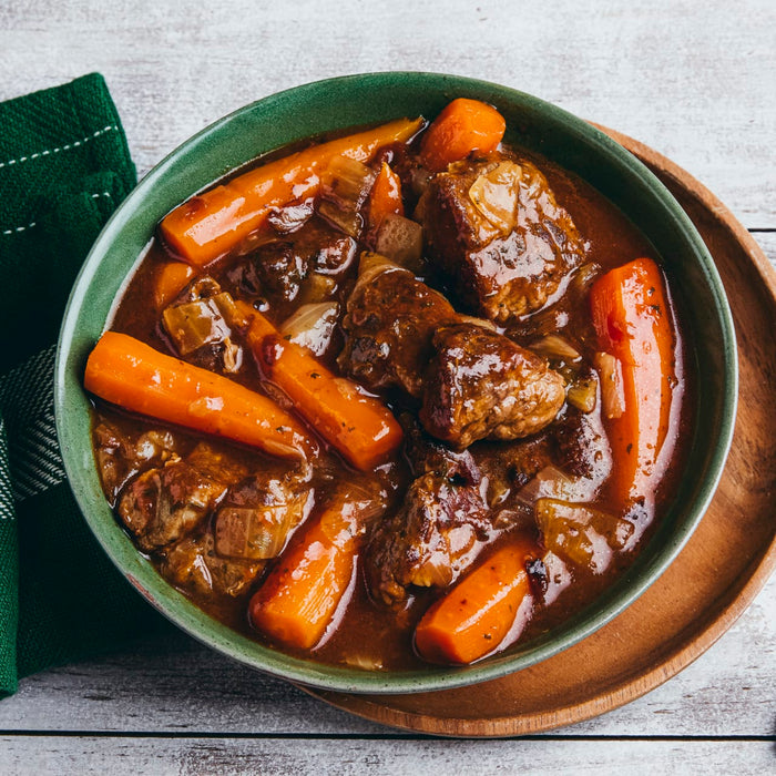 A hearty bowl of beef stew with large chunks of carrots is presented in a green bowl on a white wooden table.