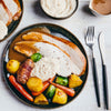 A plated roast turkey dinner with gravy, roasted potatoes, carrots, Brussels sprouts, and a sausage wrapped in bacon is set on a white wooden table next to a fork and knife.