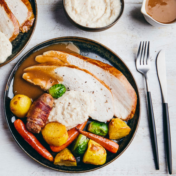A plated roast turkey dinner with gravy, roasted potatoes, carrots, Brussels sprouts, and a sausage wrapped in bacon is set on a white wooden table next to a fork and knife.