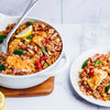 A large white serving dish is filled with a savory rice dish containing chicken, bacon, and peppers, with a portion served on a plate to the side.
