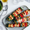 Four grilled chicken and vegetable skewers are arranged on a grey platter, served with a side salad and vinaigrette.