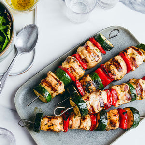 Four grilled chicken and vegetable skewers are arranged on a grey platter, served with a side salad and vinaigrette.