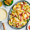 A platter of tortilla chips is generously topped with creamy cheese sauce, crispy bacon bits, and sliced jalapeños, with a bowl of extra cheese sauce on the side.