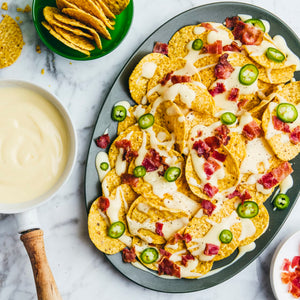 A platter of tortilla chips is generously topped with creamy cheese sauce, crispy bacon bits, and sliced jalapeños, with a bowl of extra cheese sauce on the side.