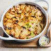 A high-angle shot of a hearty chicken, leek, and potato gratin in a white ceramic dish, with a spoonful resting on a wooden board beside it.