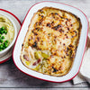 A freshly baked chicken and leek casserole with a golden-brown cheesy top, served in a red-rimmed baking dish next to a bowl of mashed potatoes and peas.