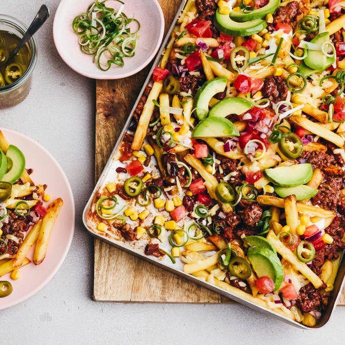 A large tray of loaded chili cheese fries is seen from above, topped with avocado, jalapeños, and corn, with a portion served on a pink plate.