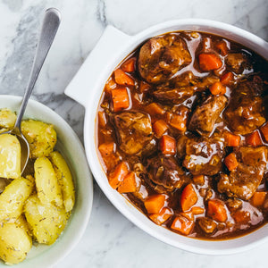 A white casserole dish filled with a rich meat and carrot stew sits on a marble countertop next to a bowl of herb-seasoned boiled potatoes.
