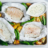 A top-down view of a sheet pan dinner featuring three portions of fish cooked in parchment paper, served with roasted potatoes, green snap peas, and a creamy herb sauce in a small gravy boat.