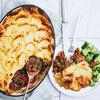 A freshly baked shepherd's pie with a golden-brown mashed potato topping is shown in a baking dish, with a serving on a white plate next to it accompanied by broccoli.