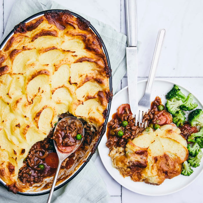 A freshly baked shepherd's pie with a golden-brown mashed potato topping is shown in a baking dish, with a serving on a white plate next to it accompanied by broccoli.