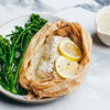 A fillet of fish cooked in parchment paper is served on a plate with a creamy sauce, lemon slices, and a side of broccolini.