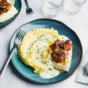 A beautifully plated meal features a fillet of white fish and a swirl of mashed potatoes, all drizzled with a creamy herb sauce and topped with crispy bacon.