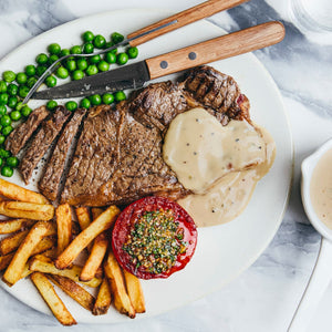 An overhead shot of a plated meal featuring sliced steak with a creamy sauce, french fries, green peas, and a grilled tomato.