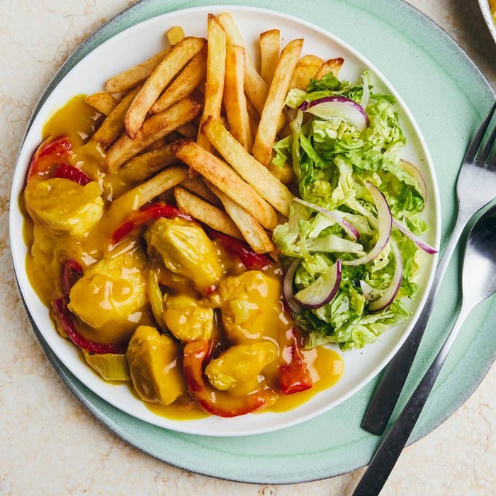 A top-down view of a white plate featuring chicken curry, a pile of golden french fries, and a side salad with red onion.