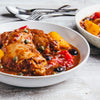 A close-up of a white bowl filled with chicken cacciatore, featuring tender chicken thighs, red and yellow peppers, black olives, and capers in a savory tomato sauce, all served on a rustic white wooden table.