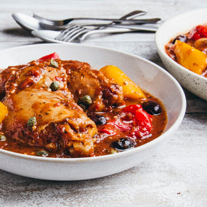 A close-up of a white bowl filled with chicken cacciatore, featuring tender chicken thighs, red and yellow peppers, black olives, and capers in a savory tomato sauce, all served on a rustic white wooden table.