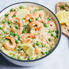 A white bowl with a black rim is filled with a creamy shrimp and pea risotto, garnished with fresh dill and lemon slices.