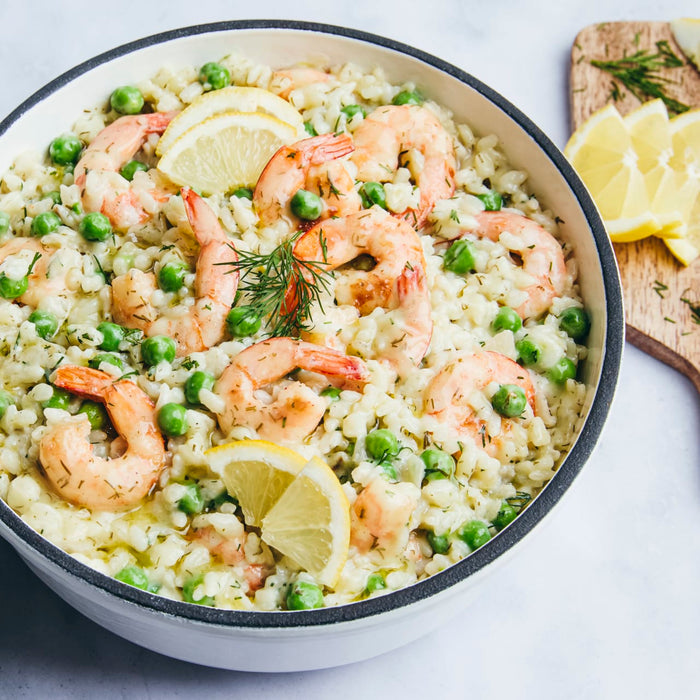 A white bowl with a black rim is filled with a creamy shrimp and pea risotto, garnished with fresh dill and lemon slices.