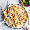 A top-down view of a creamy spaghetti carbonara with bacon and mushrooms served in a white skillet, with a side salad of fresh greens.