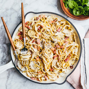 A top-down view of a creamy spaghetti carbonara with bacon and mushrooms served in a white skillet, with a side salad of fresh greens.