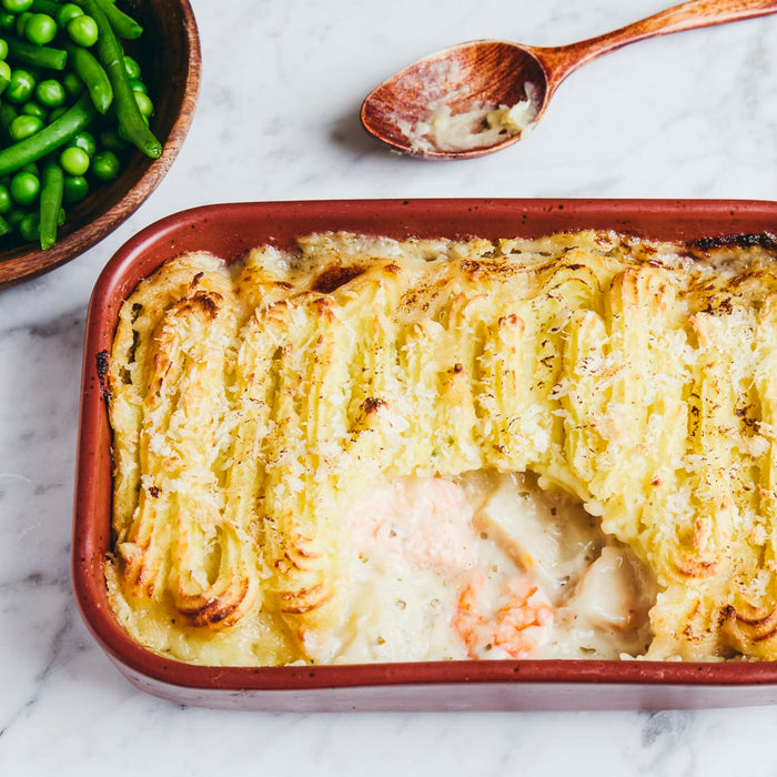 A freshly baked fish pie in a terracotta dish, with a golden-brown mashed potato topping, is served on a marble surface next to a bowl of green peas.