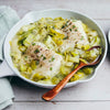 A white serving dish contains two pieces of poached fish nestled in a creamy leek sauce, garnished with fresh herbs and black pepper.