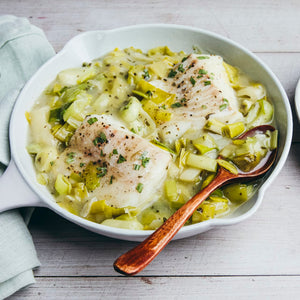 A white serving dish contains two pieces of poached fish nestled in a creamy leek sauce, garnished with fresh herbs and black pepper.