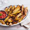 A silver basket filled with seasoned potato wedges sits on a white wooden table, accompanied by a small bowl of red dipping sauce.