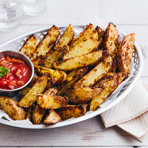 A silver basket filled with seasoned potato wedges sits on a white wooden table, accompanied by a small bowl of red dipping sauce.